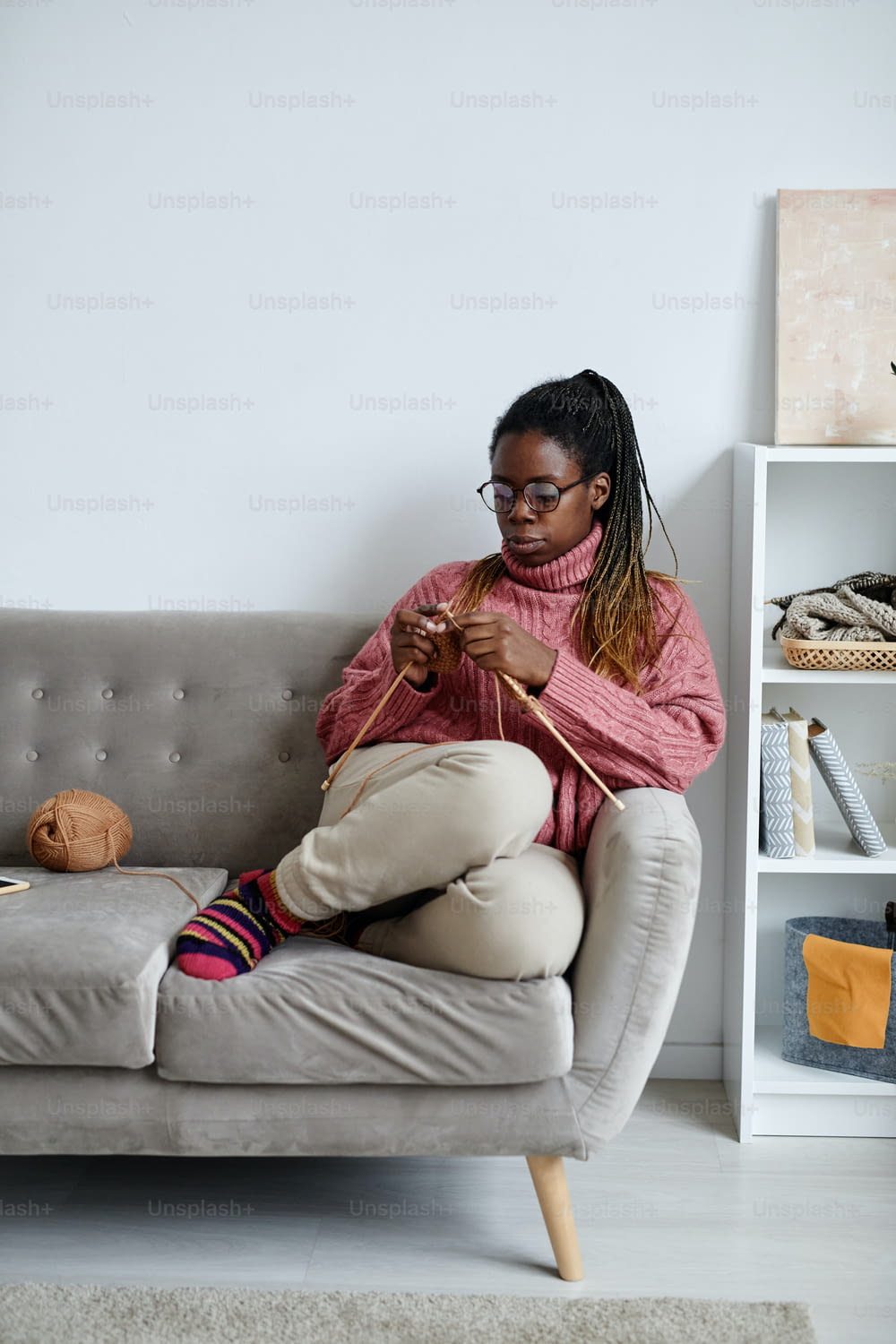 Vertikales Ganzkörperporträt einer jungen afroamerikanischen Frau, die strickt, während sie ein gemütliches Wochenende zu Hause genießt