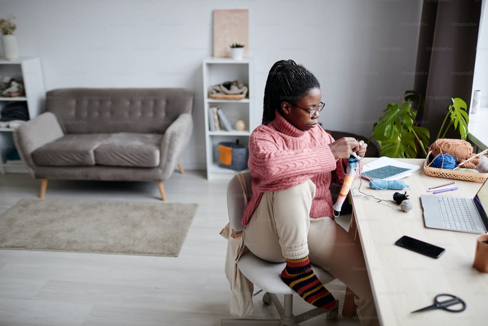 High angle portrait of young African-American woman knitting at home and looking at laptop screen, watching educational videos, copy space