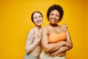 Cintura para cima retrato de duas mulheres jovens sorridentes pele escura e pele clara abraçando enquanto em pé contra fundo amarelo vibrante em roupas íntimas