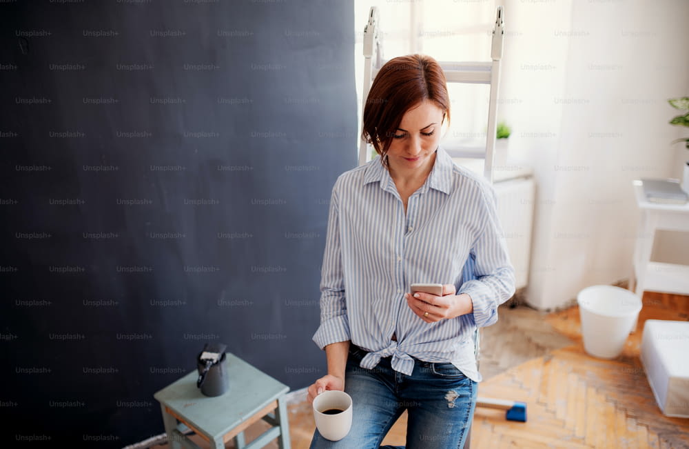 A portrait of young creative woman with a cup of coffee and smartphone painting wall black. A startup of small business.