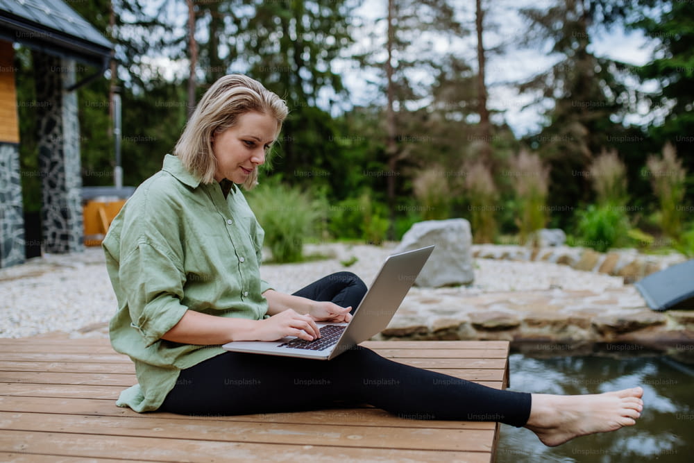 Uma mulher, freelancer trabalhando em laptop, sentada em um píer à beira do lago do quintal, um conceito de escritório remoto, trabalho durante as férias.