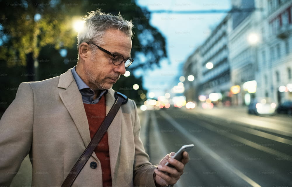 Ein reifer Geschäftsmann mit Smartphone, der abends auf einer Straße steht und eine SMS schreibt. Speicherplatz kopieren.