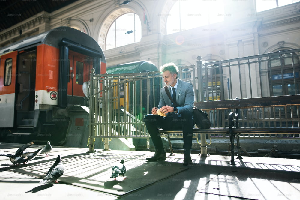 Bello uomo d'affari maturo in una città. Un uomo che aspetta il treno alla stazione ferroviaria. Un uomo seduto su una panchina, che dà da mangiare ai piccioni.