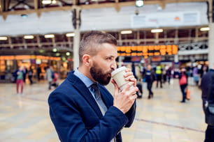 Hipster businessman holding a disposable cup and drinking coffee at the crowded train station
