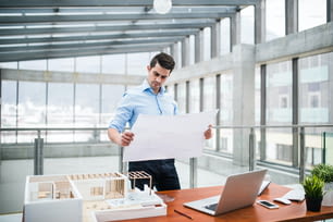 Junger Geschäftsmann oder Architekt mit Modell eines Hauses und Blaupausen, die am Schreibtisch im Büro stehen und arbeiten.