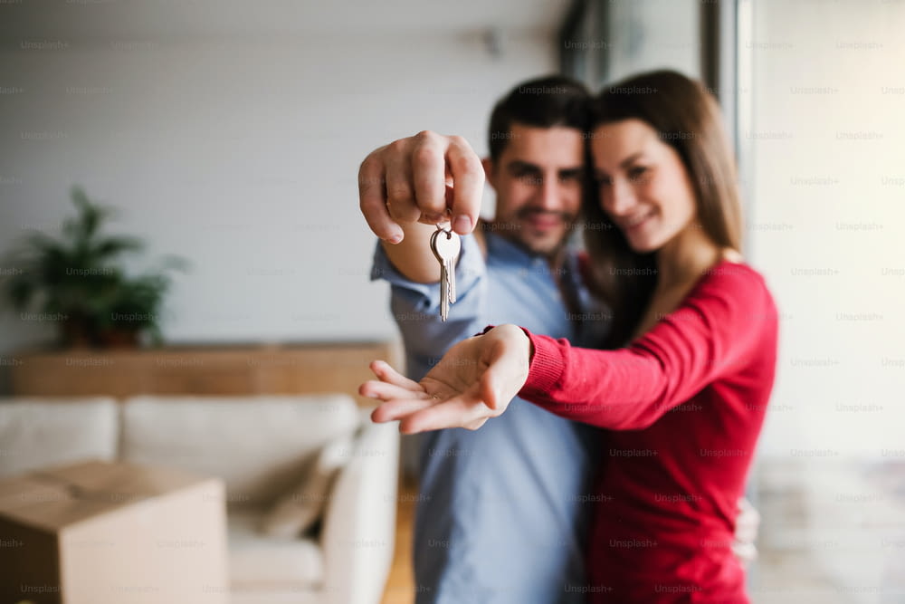 Un jeune couple heureux avec une clé et des boîtes en carton debout à l’intérieur, emménageant dans une nouvelle maison.