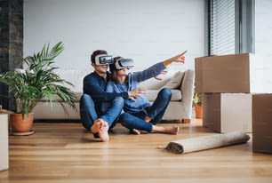 A young couple with VR goggles and cardboard boxes sitting barefoot on a floor, moving in a new home.