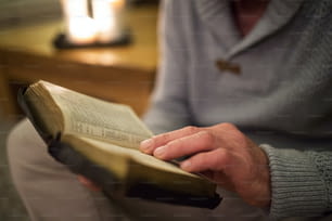 Homem idoso irreconhecível em casa em sua sala de estar, sentado no chão, lendo a Bíblia. Velas acesas atrás dele.