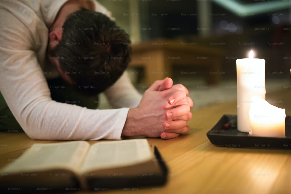 Unrecognizable young man praying, kneeling on the floor, hands clasped together. Bible and burning candles next to him. Close up.