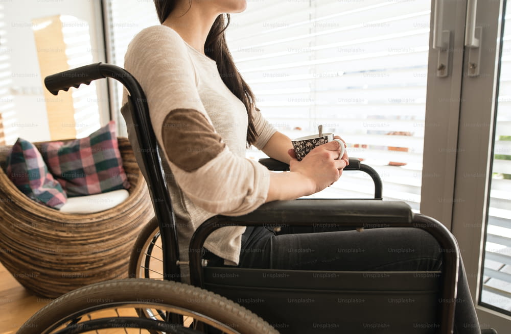 Unrecognizable young disabled woman in wheelchair at home in her living room, looking out the window, holding a cup of tea or coffee