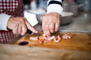 Unrecognizable senior woman cooking at home. Close up of hands chopping bacon.