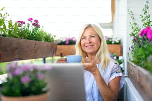 Mujer mayor activa con computadora portátil y café trabajando en balcón, concepto de oficina en casa.