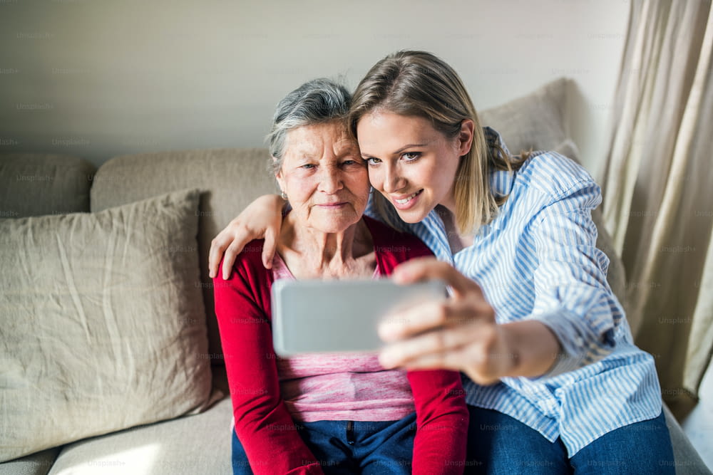 An elderly grandmother and adult granddaughter with smartphone at home, sitting on a sofa and taking selfie.