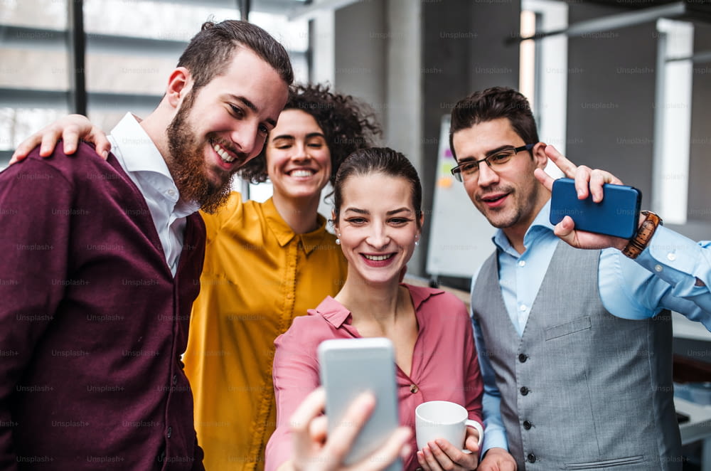 A group of cheerful young businesspeople with smartphone in office, taking selfie.
