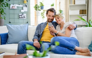 Front view of happy couple in love sitting indoors at home, using smartphone.
