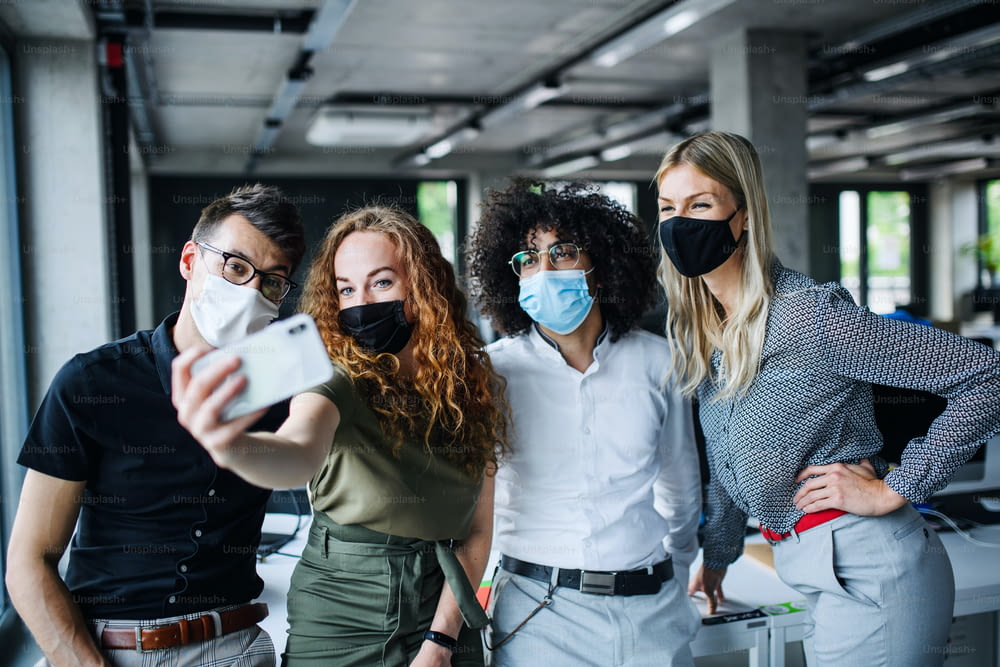 Les jeunes avec des masques faciaux de retour au travail au bureau après la quarantaine et le confinement liés au coronavirus, prenant des selfies.