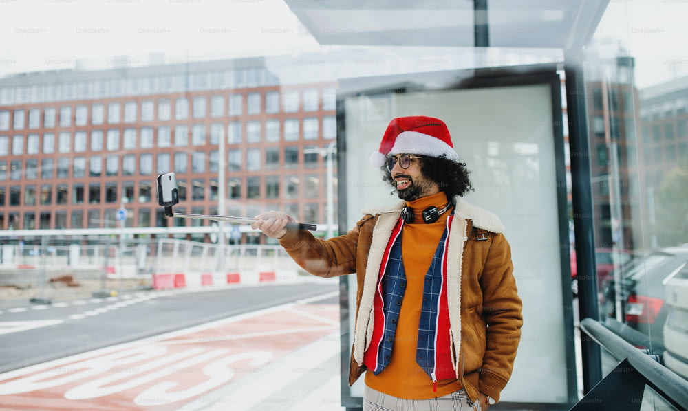 Joven con sombrero de Santa haciendo videos para las redes sociales al aire libre en una parada de autobús. Espacio de copia.