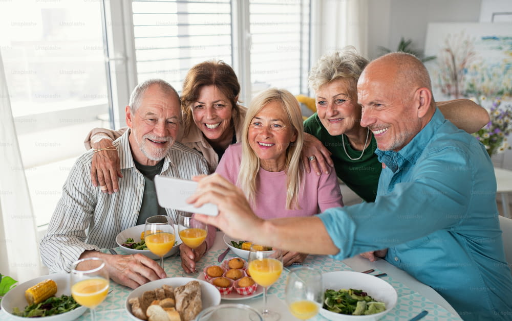 Eine Gruppe älterer Freunde, die drinnen feiern und beim Essen am Tisch ein Selfie machen.
