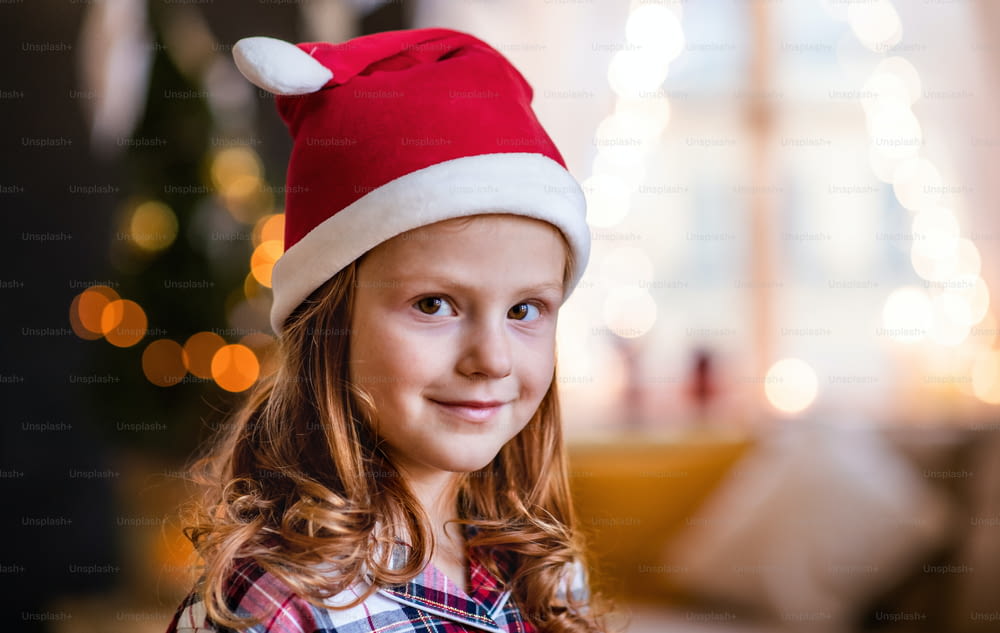 Close-up portrait of small girl with Santa hat indoors at home at Christmas, looking a camera.
