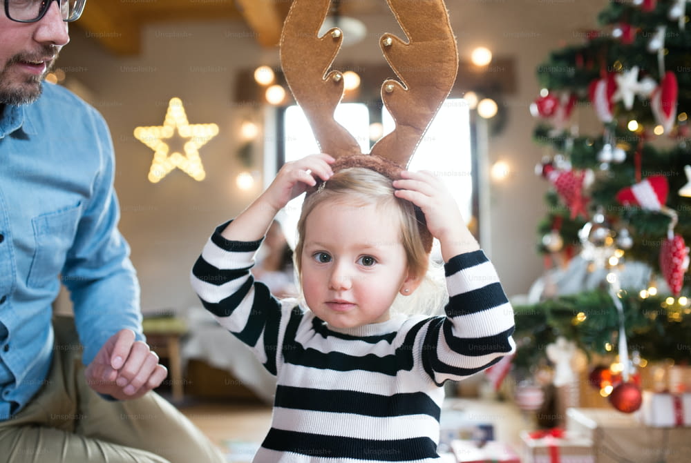 Padre joven con su linda hijita en el árbol de Navidad, vistiendo astas de reno marrón.