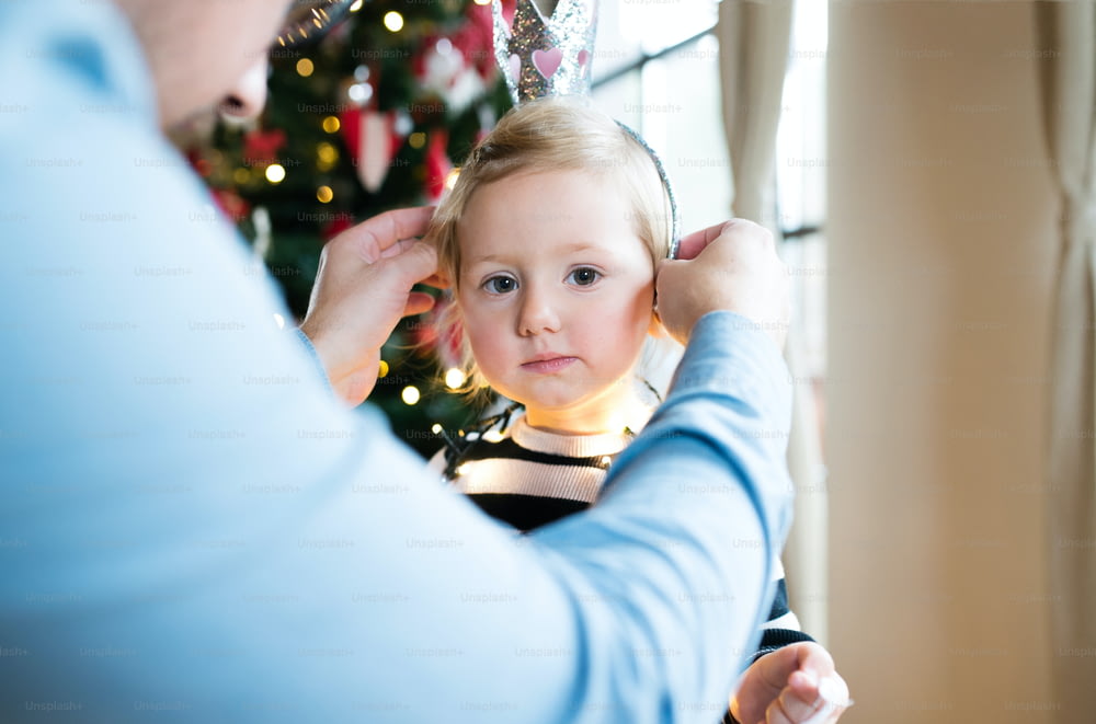Unrecognizable father at Christmas tree giving her cute little daughter crown.