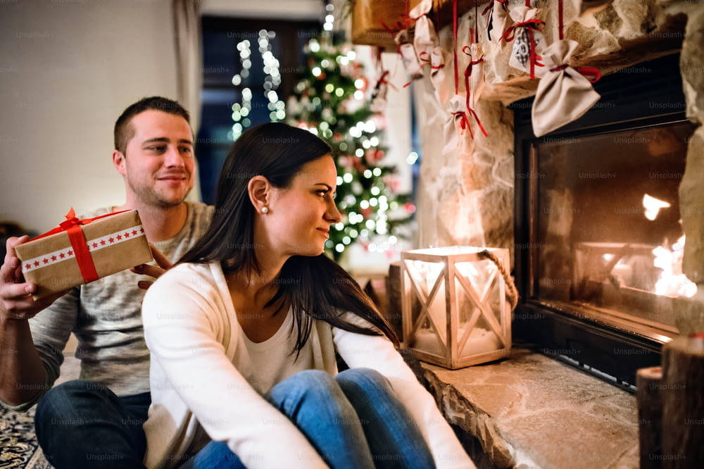 暖炉の前の床に座っている美しい若い夫婦。美しい女性にクリスマスプレゼントを贈る男性。