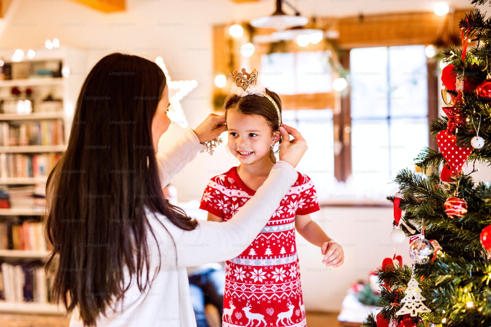 Beautiful young mother with little daughter at Christmas tree at home. Mother putting a headband on her daughters head.