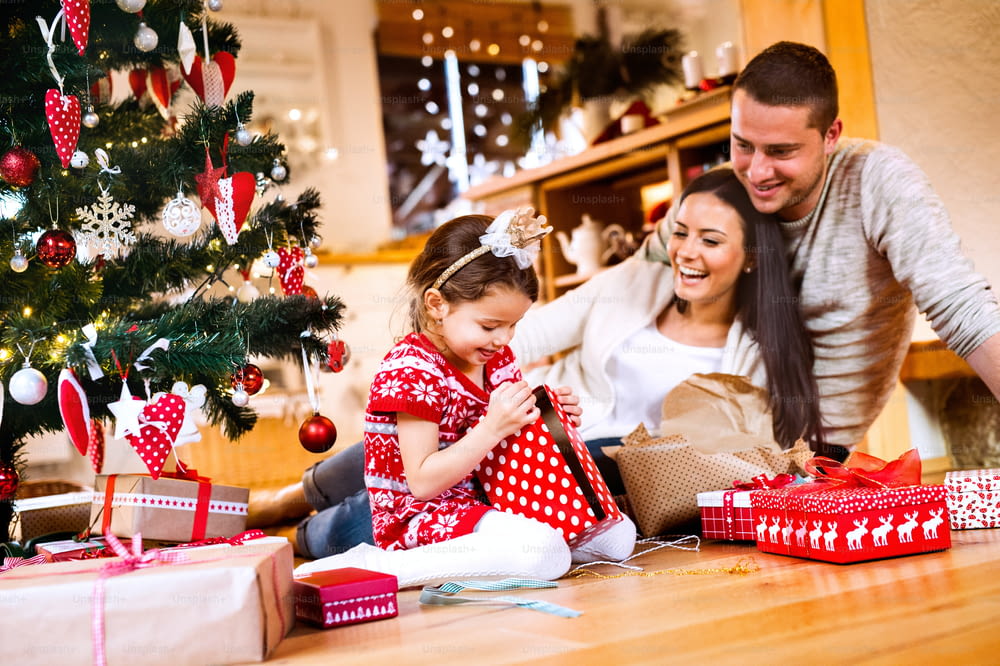 Beautiful young family with little daughter at Christmas tree at home unpacking presents.