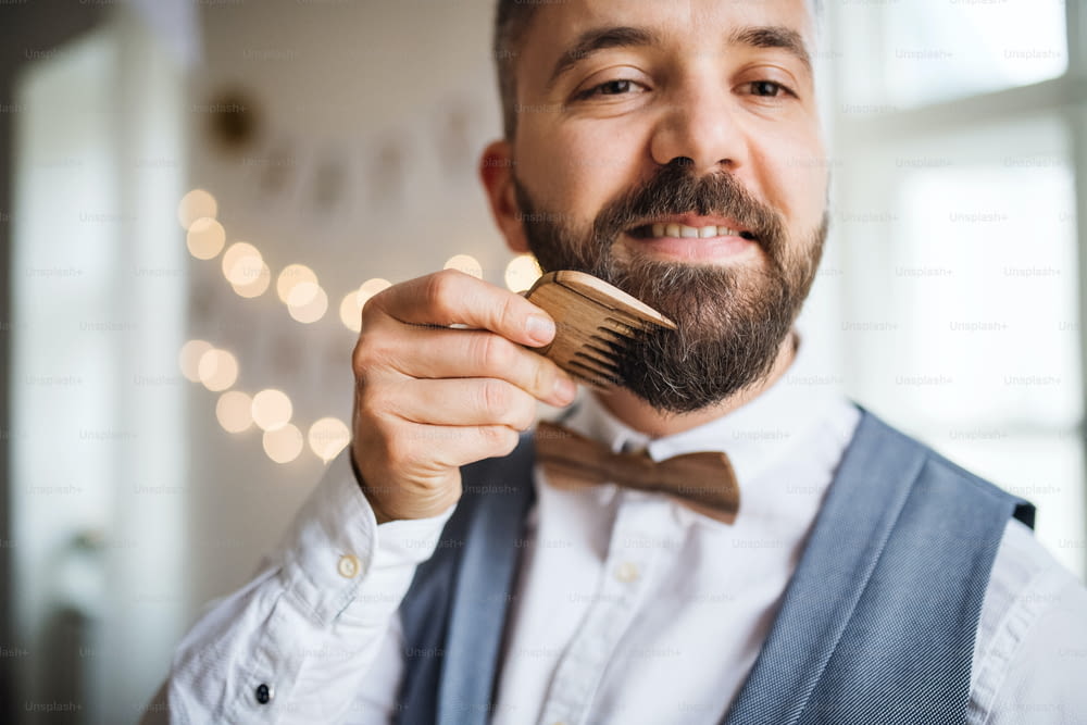 Hipster man standing indoors in a room set for a party, combing beard with a comb.