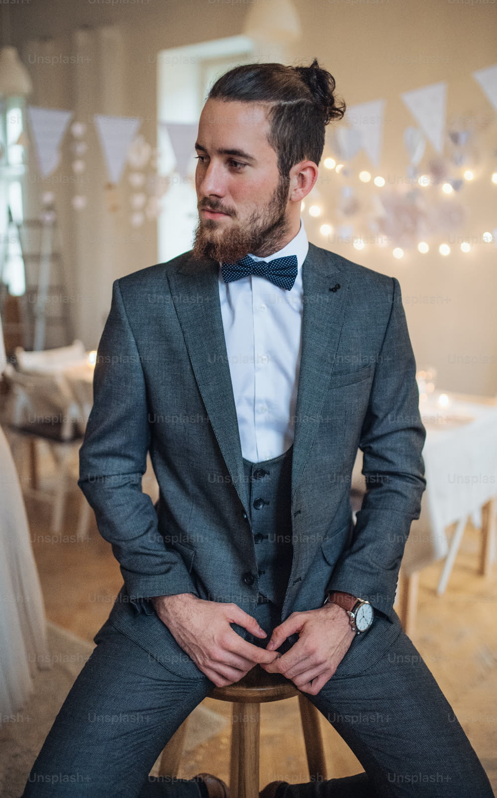 A handsome hipster young man with formal suit sitting on a stool on an indoor party, looking away.