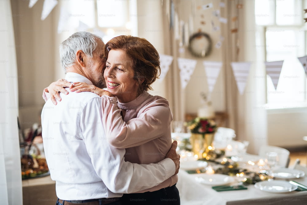 A portrait of a senior couple standing indoors in a room set for a party, hugging.