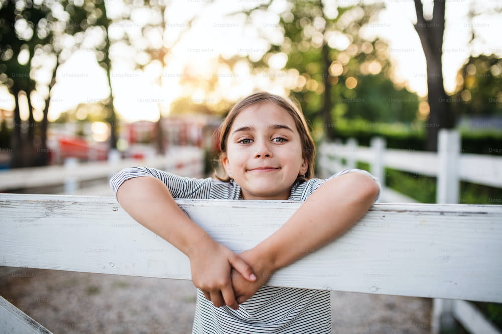 Um retrato de menina pequena feliz na fazenda da família, de pé ao lado do portão de madeira.