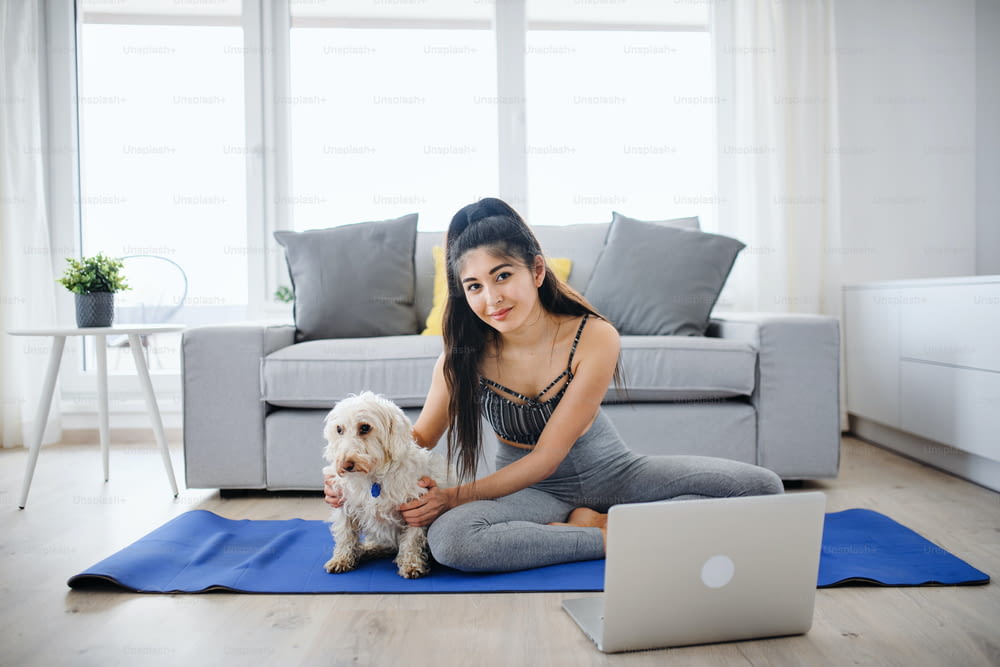 A young sport woman with laptop and dog doing exercise indoors at home, looking at camera.