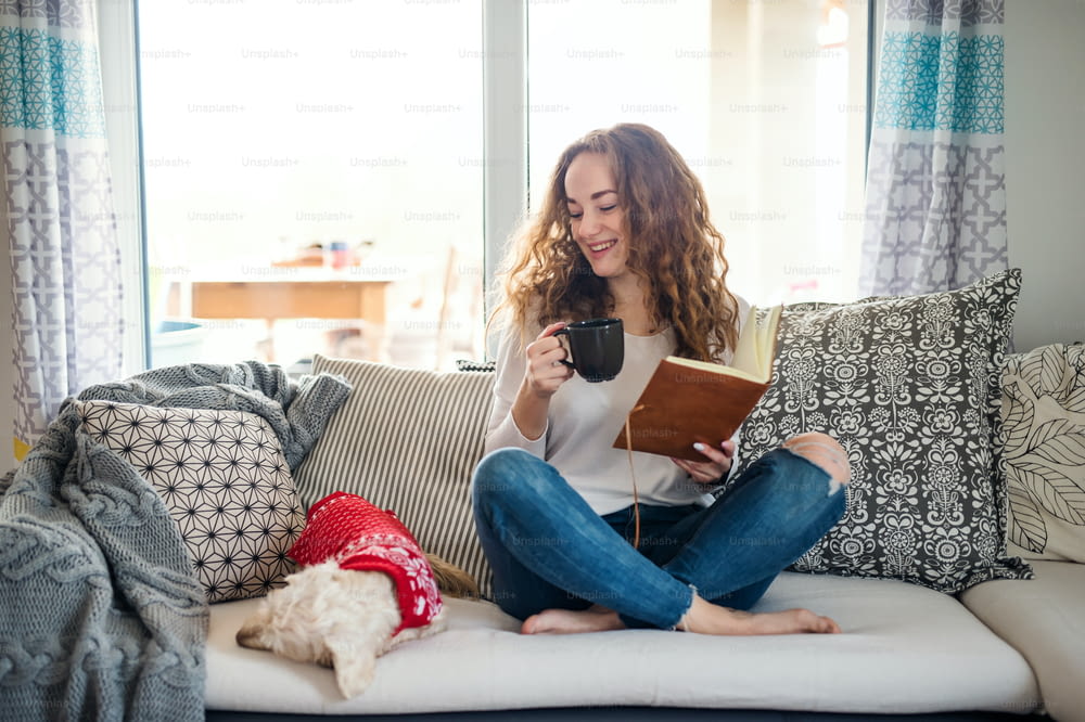 Young woman with dog and book relaxing on sofa indoors at home, reading.