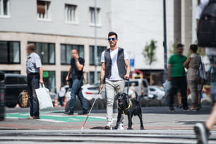 Ein junger blinder Mann mit weißem Stock und Blindenhund geht in der Stadt auf dem Bürgersteig spazieren.