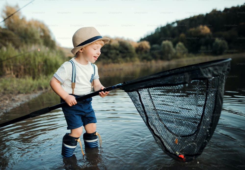 水の中に立ち、湖のほとりで網を持って釣りをしている幸せな小さな幼児の男の子。