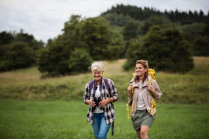 An active senior woman with binoculars hiking with her adult daughter outdoors in nature.A happy mid adult woman with trekking poles hiking with active senior mother outdoors in nature.
