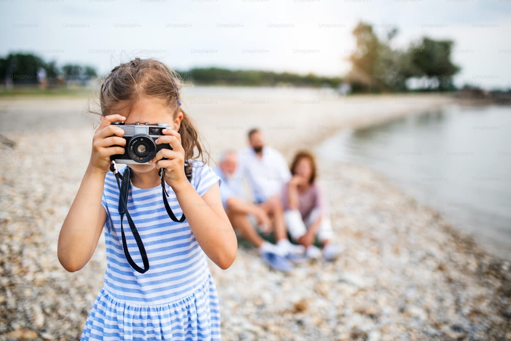 Front view of small girl with camera on a holiday with family by the lake, taking photos.