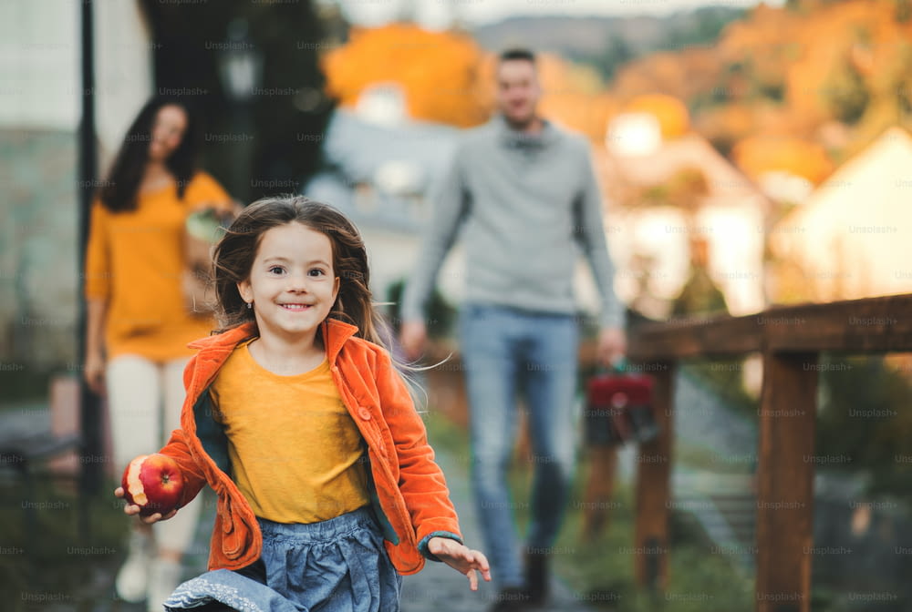 Niña pequeña con padres irreconocibles en el fondo corriendo en el parque en otoño, comiendo manzana.