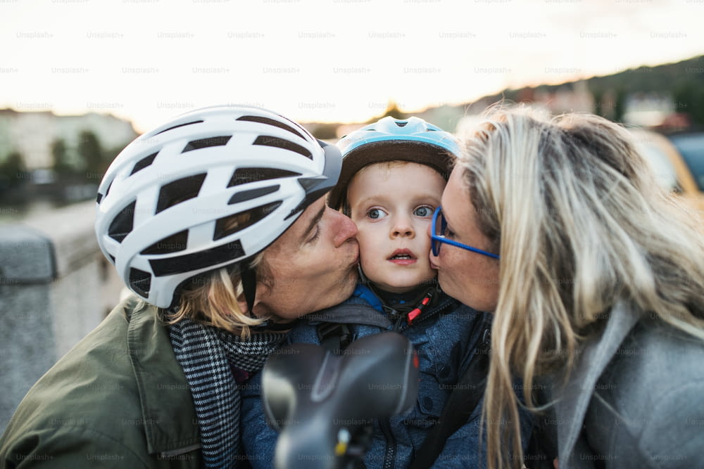 자전거 헬멧을 쓴 작은 소년과 도시의 야외에서 젊은 부모들이 키스를 하고 있다.