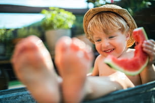 여름에 정원에 있는 야외 욕조에서 모자를 쓰고 수박을 먹는 행복한 작은 소년.