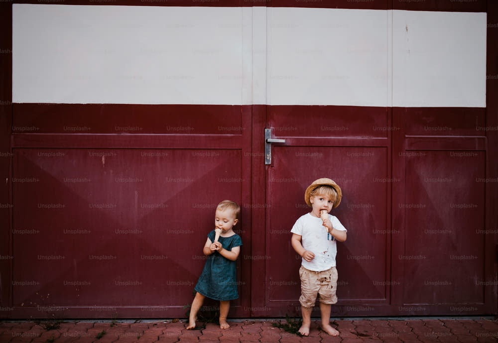 Two small toddler children outdoors in summer, eating ice cream and standing in front of a gate.