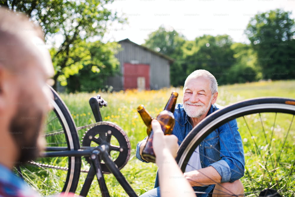 A senior father and unrecognizable son repairing bicycle outside on a sunny day, clinking glass bottles of beer.