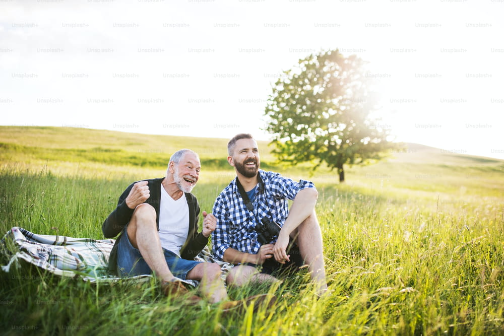 Um filho hipster adulto com seu pai mais velho sentado na grama ao pôr do sol na natureza.