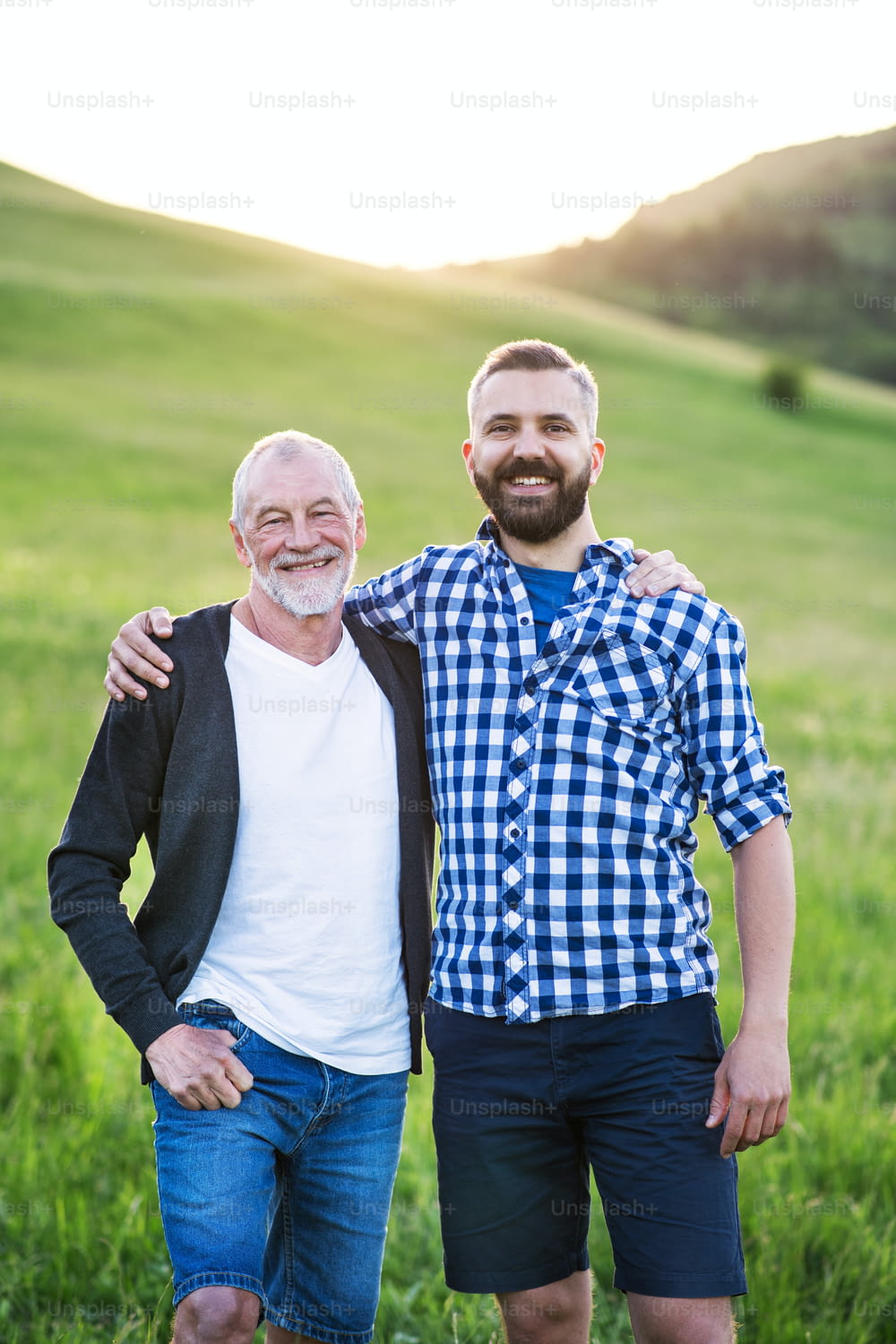 A portrait of a laughing adult hipster son with senior father in nature at sunset, arms around each other.