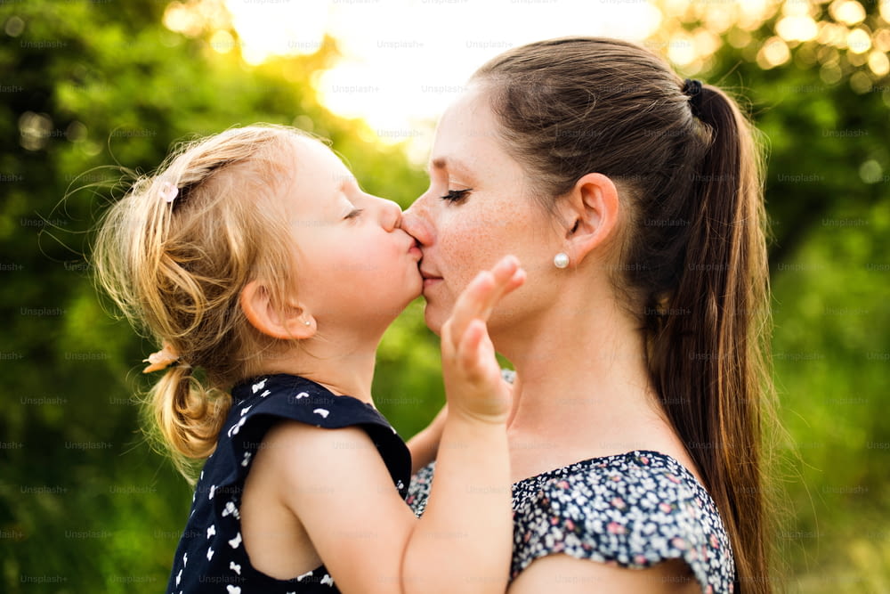 Bela jovem mãe na natureza verde ensolarada do verão segurando sua filhinha fofa nos braços, menina beijando-a no nariz.