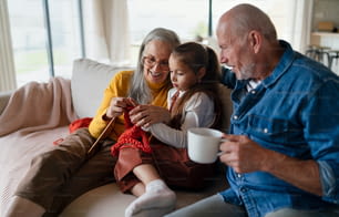 Uma garotinha sentada no sofá com seus avós e aprendendo a tricotar dentro de casa em casa.