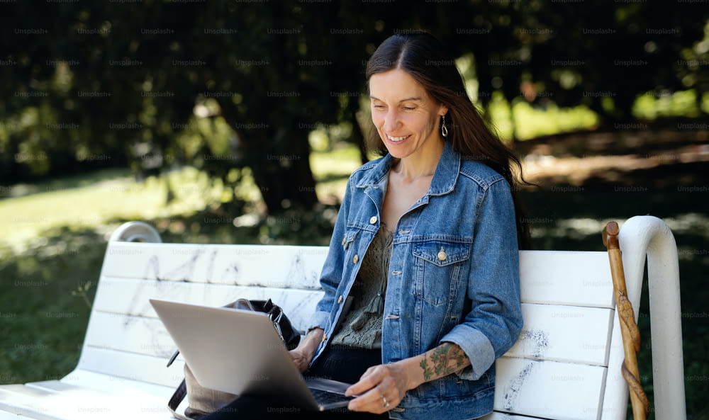 Mature woman with laptop sitting on bench outdoors in city or town park, working.