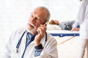 Médecin principal avec smartphone dans son bureau, faisant un appel téléphonique. Une infirmière et un patient couché à l’arrière-plan.