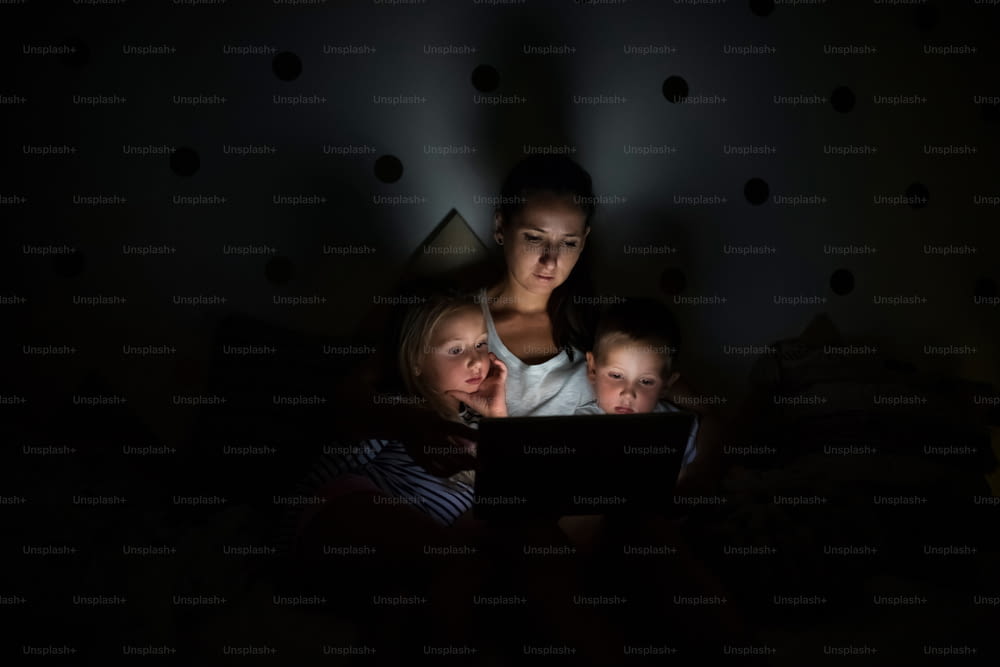 Jeune mère avec enfants assise à l’intérieur dans la chambre le soir, en utilisant un ordinateur portable.